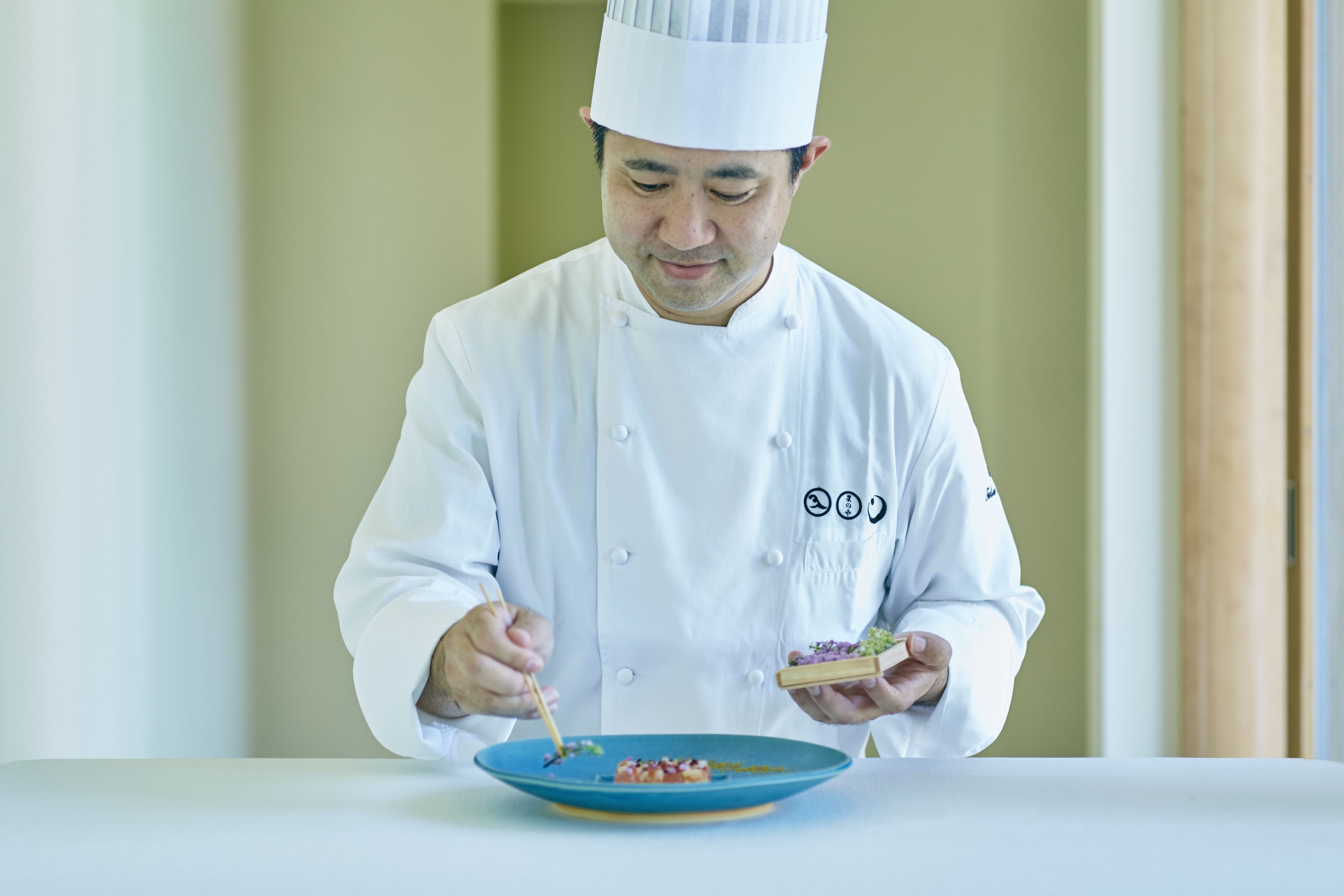 A chef in a white uniform with a hat, holding a small dish and a pair of chopsticks, standing at a table with a blue plate of food.