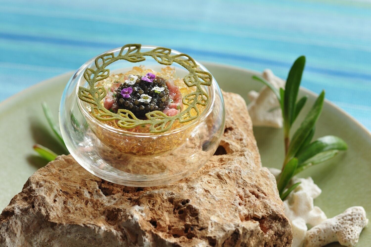 A clear glass bowl with a decorative design, placed on a textured surface with a green plate and some greenery in the background.