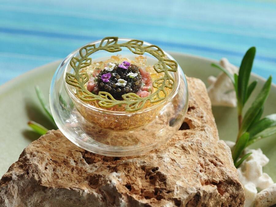 A clear glass bowl with a decorative design, placed on a textured surface with a green plate and some greenery in the background.