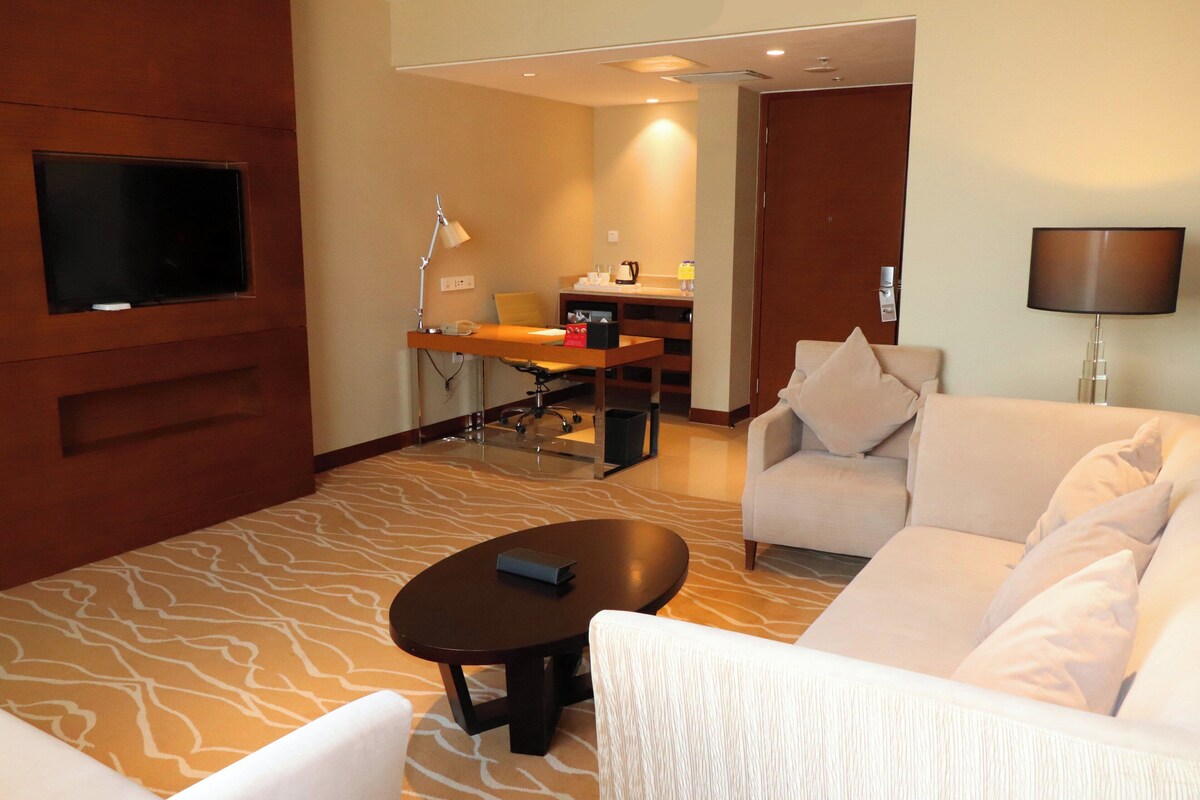 Habitación de hotel con sofá, mesa de centro, televisor montado en la pared, escritorio con silla y lámpara.