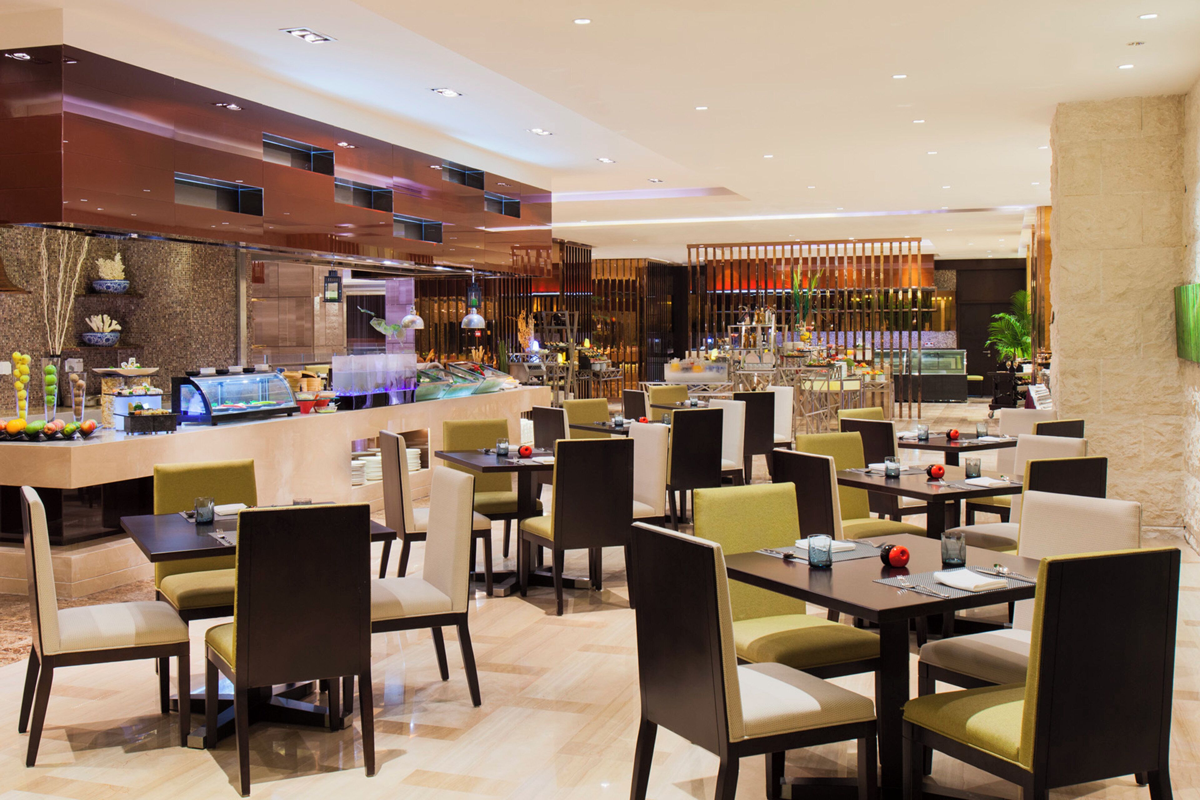 Un restaurant moderne doté de nombreuses tables et chaises, d’un bar où sont exposés divers plats, et d’un mur décoratif agrémenté d’éléments verticaux.