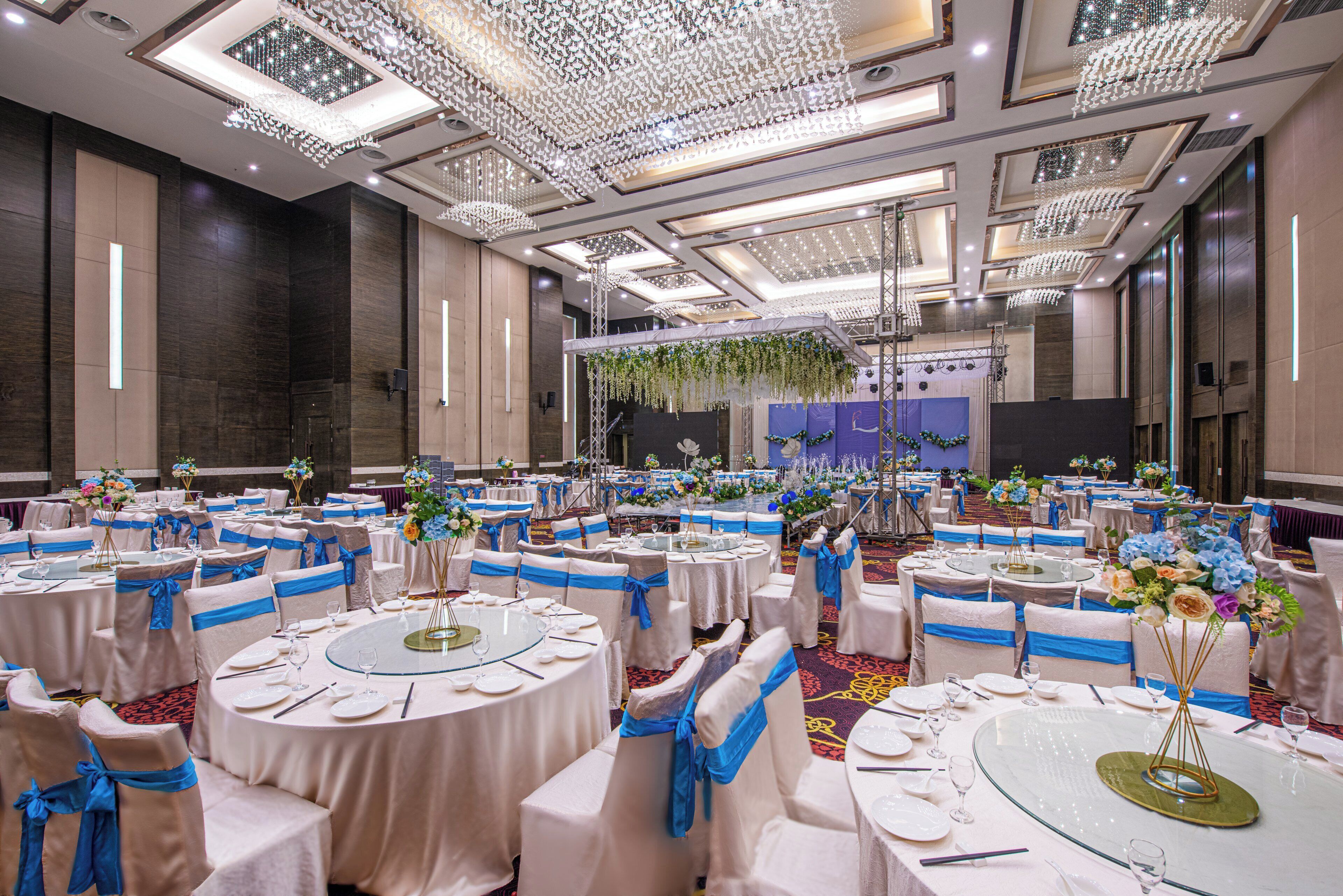 A banquet hall with round tables, chairs with blue sashes, and a stage with floral decorations.