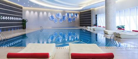 A modern indoor swimming pool with lounge chairs, a large decorative wall, and a ceiling with recessed lighting.