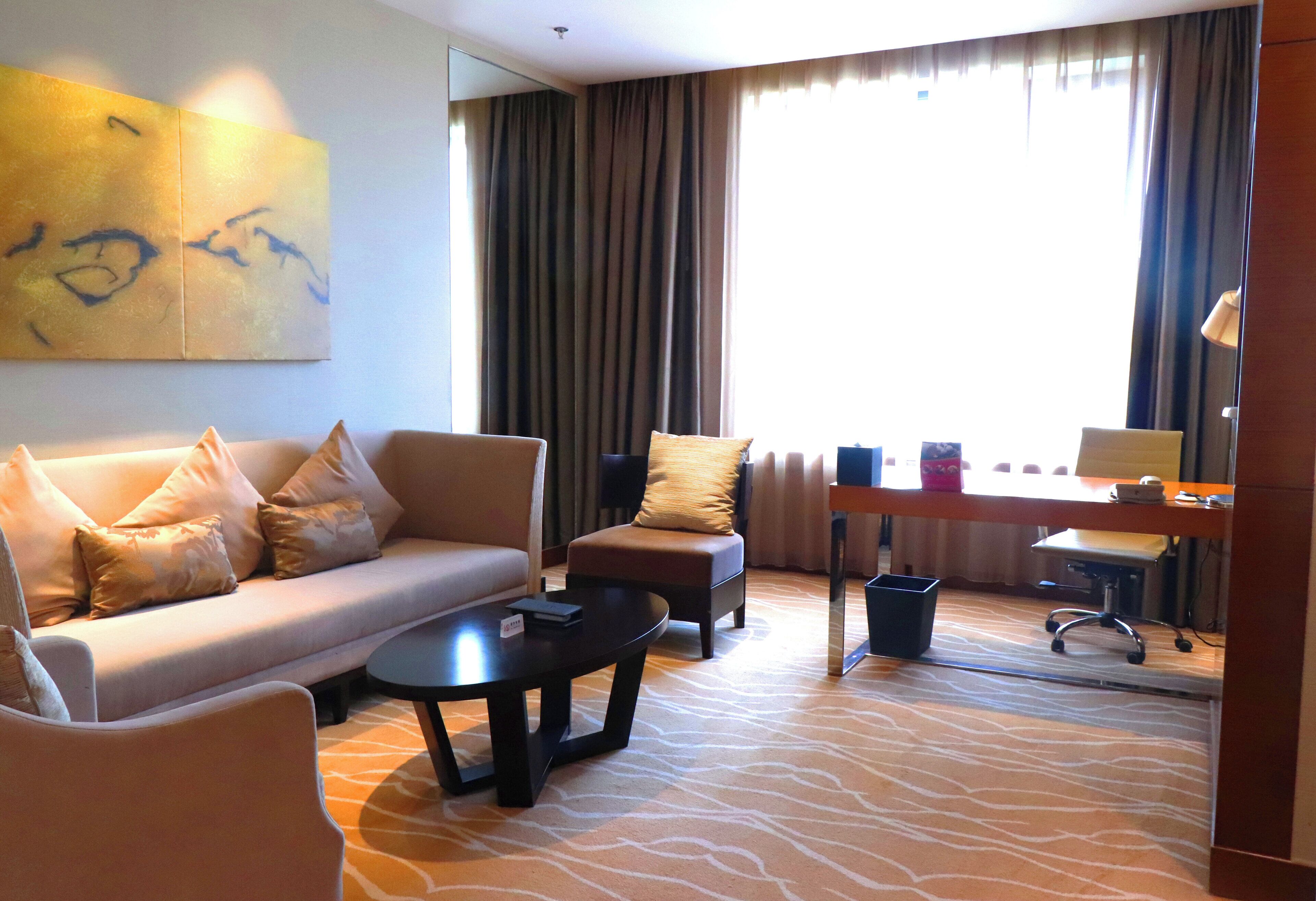 Een hotelkamer met een bank, fauteuil, salontafel, bureau, stoel en grote ramen met gordijnen.