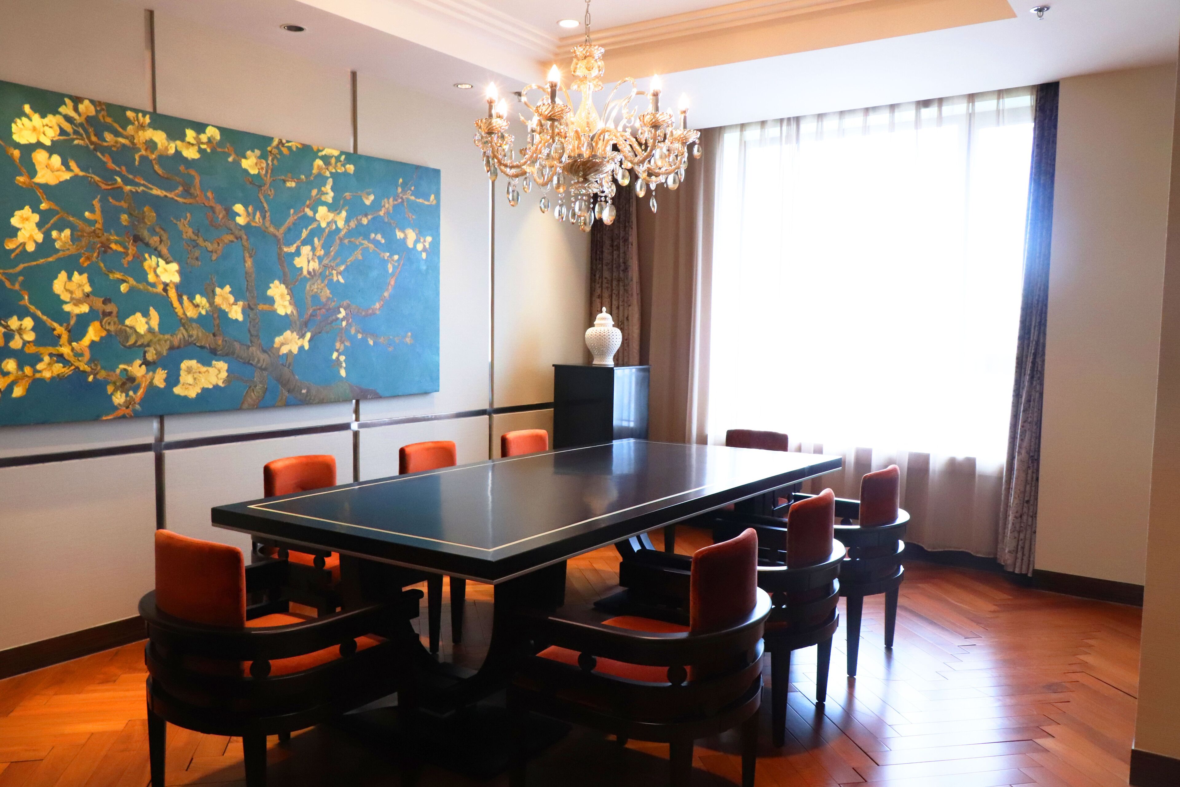 Une salle de conférence avec une longue table, six chaises, un lustre, un grand tableau et une fenêtre avec des rideaux.