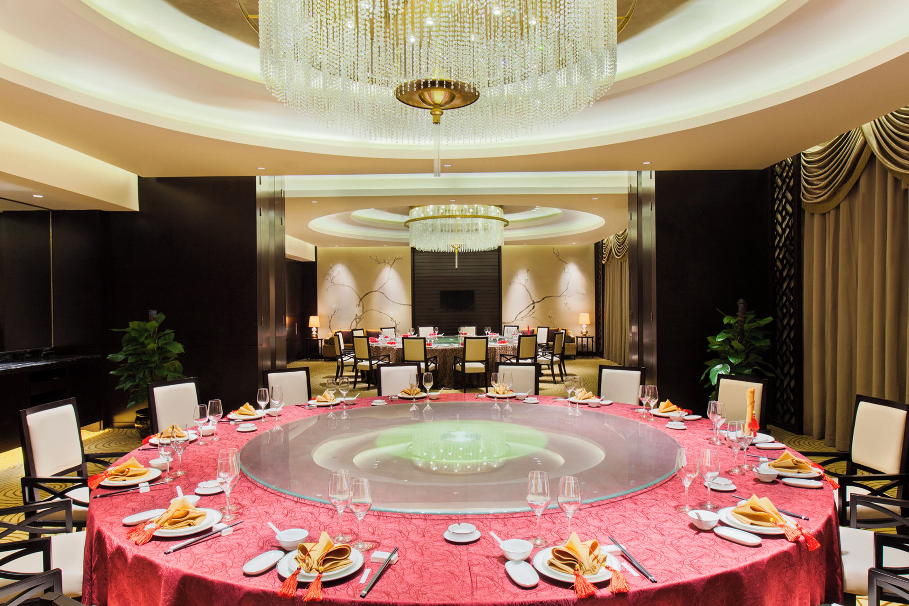 A formal dining room with a round table set for a meal, surrounded by chairs, and a large chandelier above.