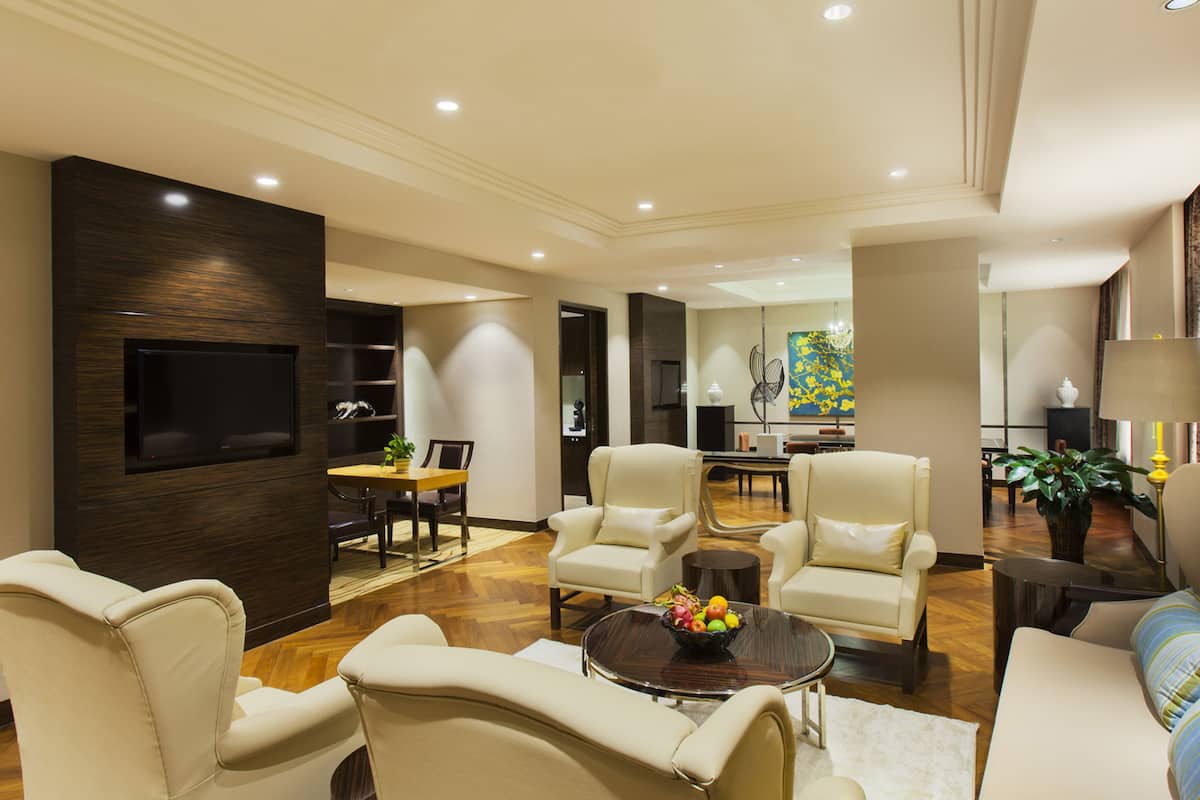 A spacious living room with a flat-screen TV mounted on a dark wood panel, a dining area with a table and chairs, and a kitchen in the background.