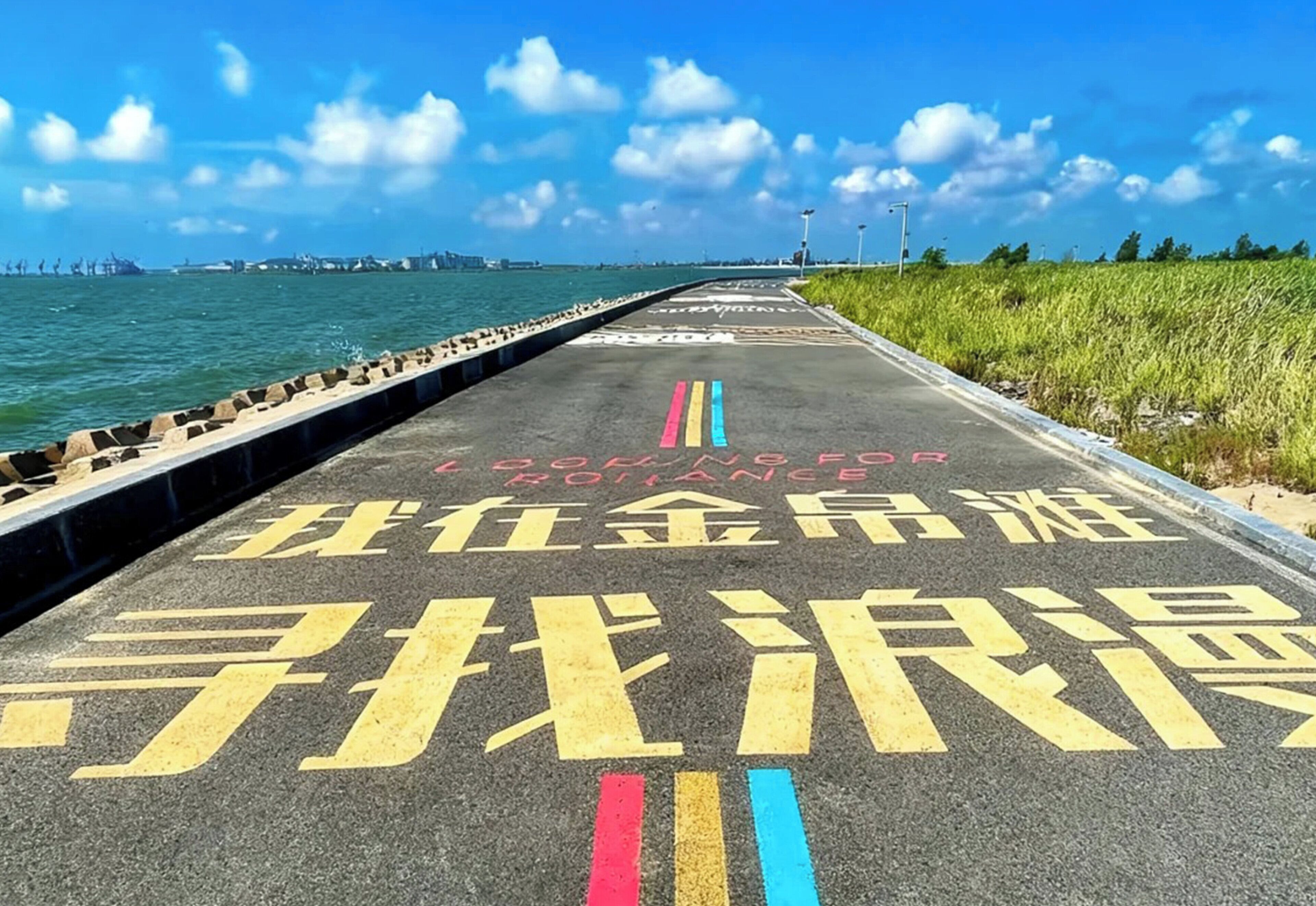 Una carretera costera con texto y símbolos pintados, que conduce a una masa de agua con estructuras industriales al fondo.
