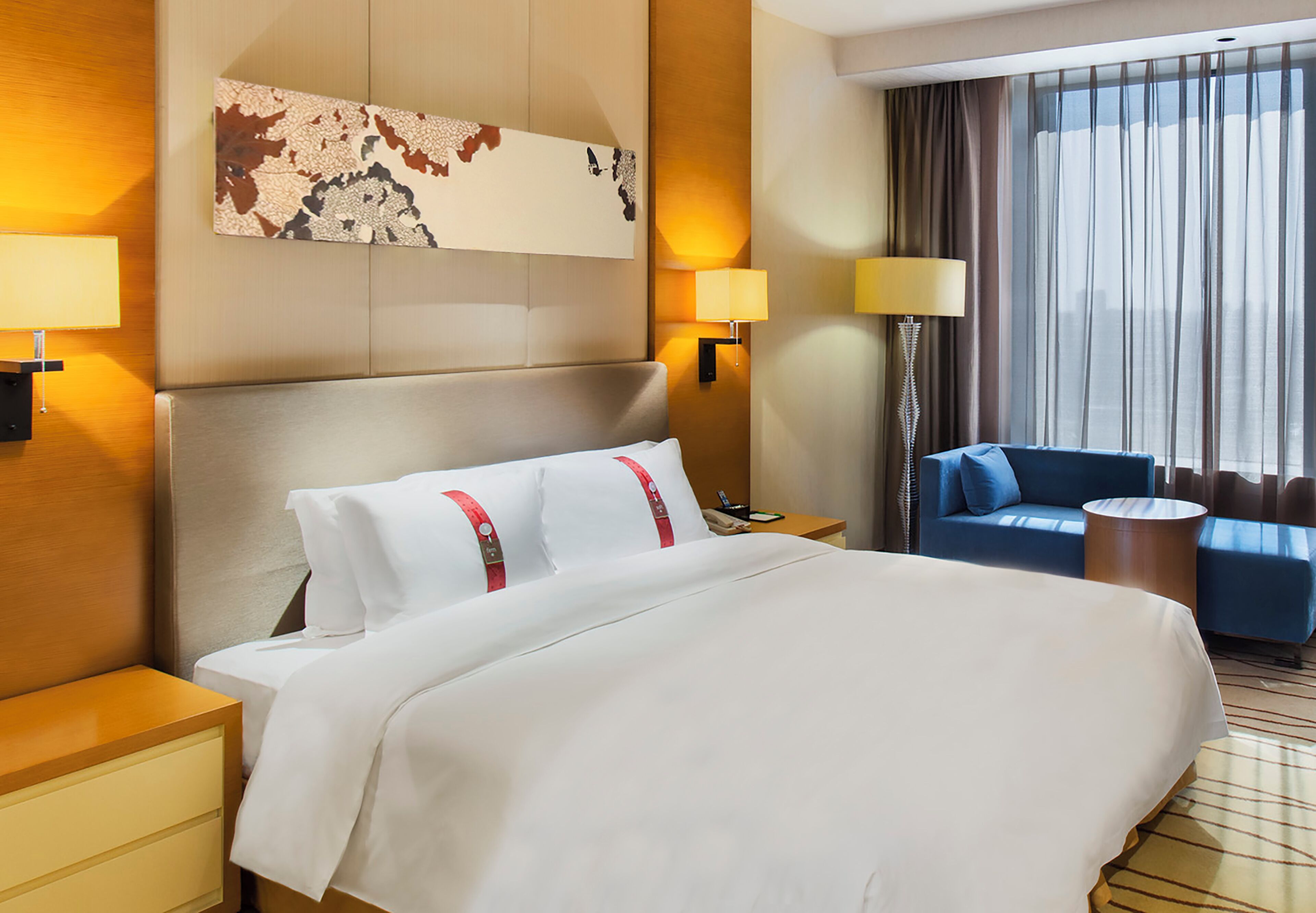 Habitación de hotel con una cama grande, dos mesitas de noche, un escritorio, una silla, un sofá y un ventanal con cortinas.
