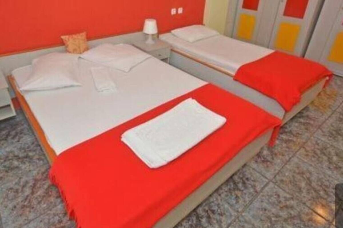 Een kamer met twee bedden, elk voorzien van witte lakens en rode dekens. Er is een nachtkastje met een lamp en een kleine tafel met een stoel. De muren zijn rood geschilderd, en er is een deur met gele en rode panelen.