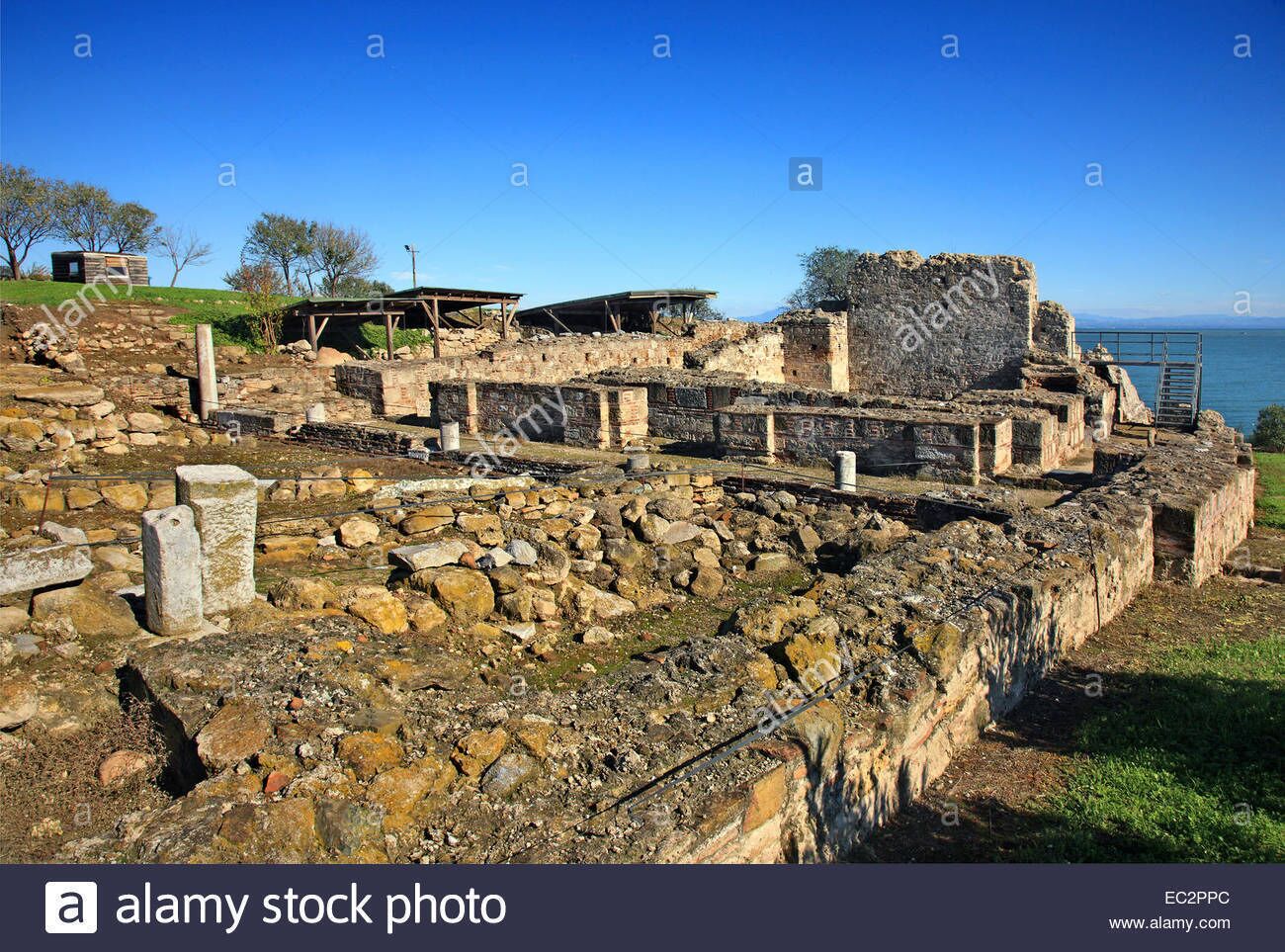 Ruínas antigas com paredes de pedra e pedras espalhadas, um portão de metal e céu claro.