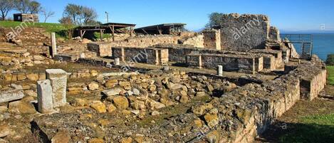 Ruinas antiguas con muros de piedra y piedras dispersas, una reja de metal y un cielo despejado.