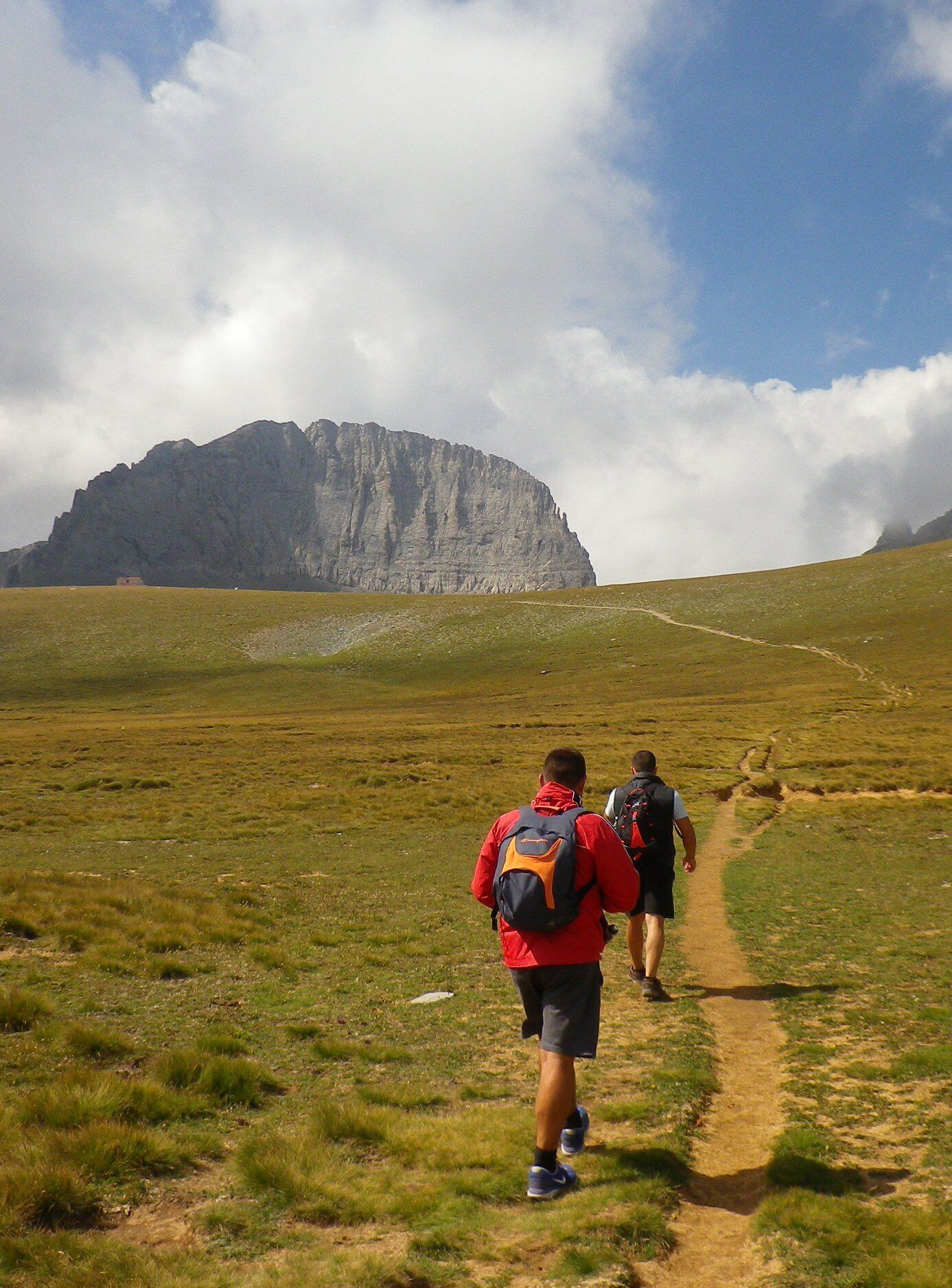 Dos excursionistas en un sendero cubierto de hierba, con una montaña al fondo.