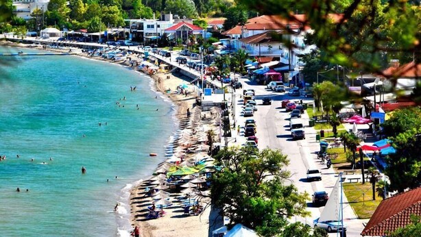 A coastal town with a beach lined with umbrellas and boats, a road with parked cars, and buildings with red roofs.