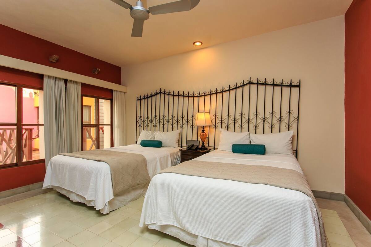 A hotel room with two beds, a ceiling fan, a window with curtains, and a decorative metal headboard.