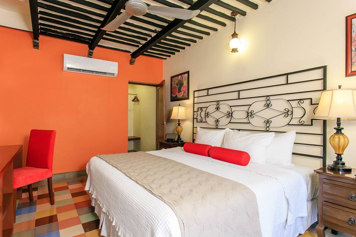 A hotel room with a bed, a red chair, a desk, a nightstand with a lamp, and a ceiling fan.