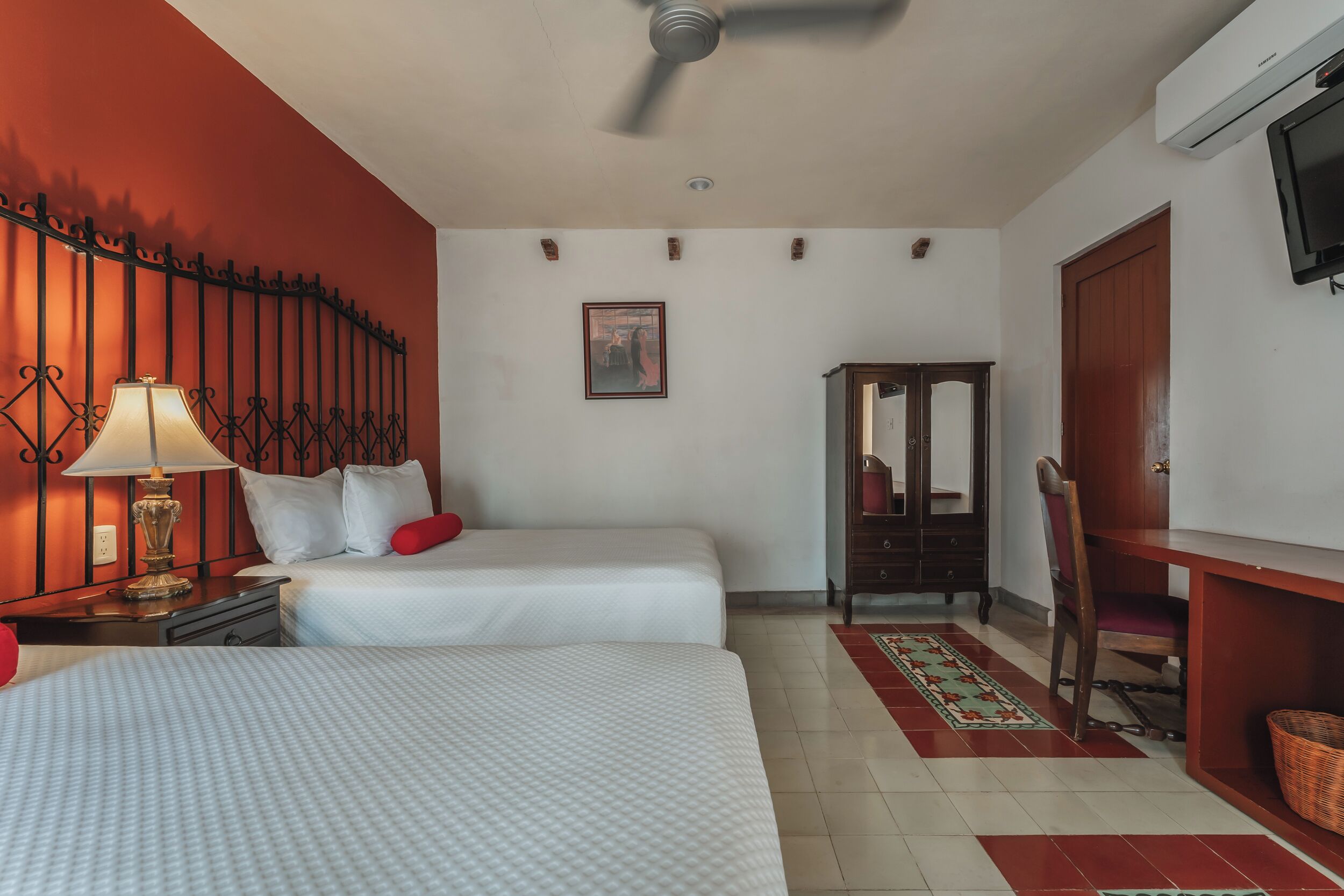 A hotel room with two beds, a desk, a chair, a television, and a ceiling fan.