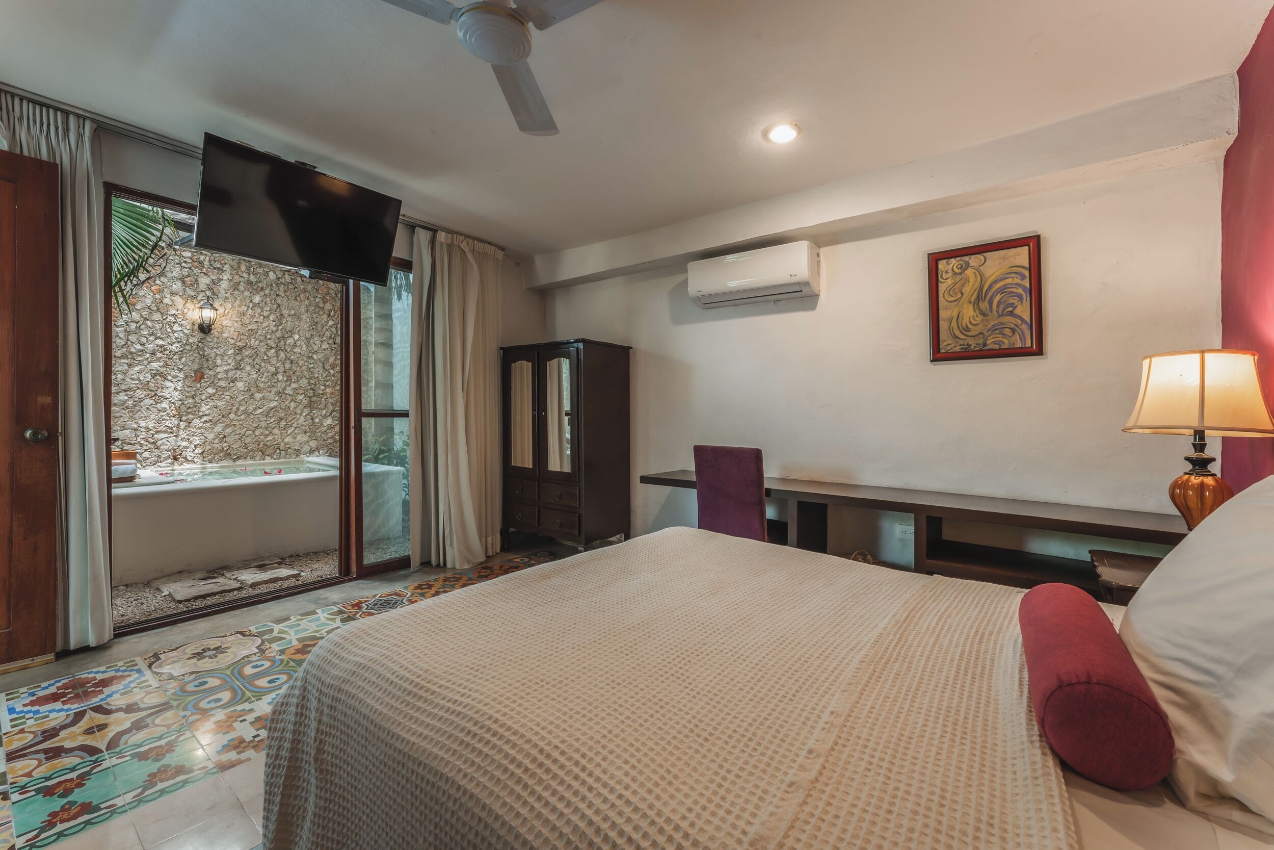 A hotel room with a bed, a desk, a chair, a television, an air conditioning unit, and a window with curtains.