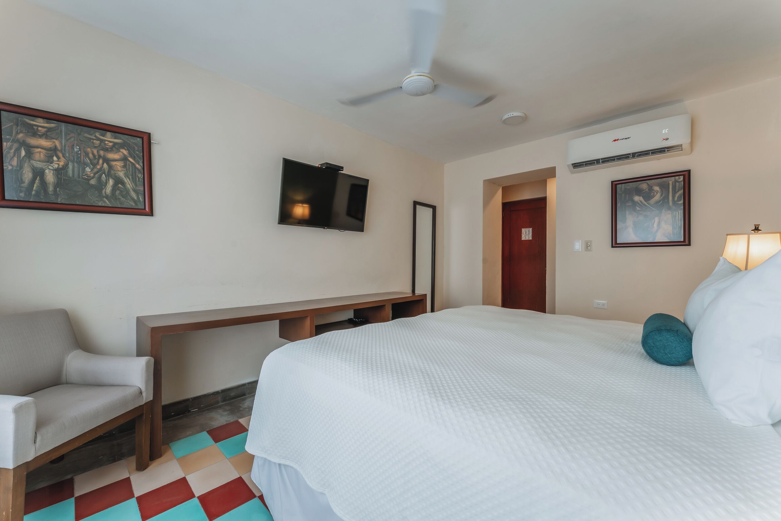 Habitación de hotel con cama, escritorio, silla, ventilador de techo y aire acondicionado, además de obras de arte en las paredes.