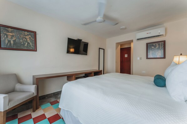 A hotel room with a bed, a desk, a chair, a ceiling fan, an air conditioning unit, and artwork on the walls.