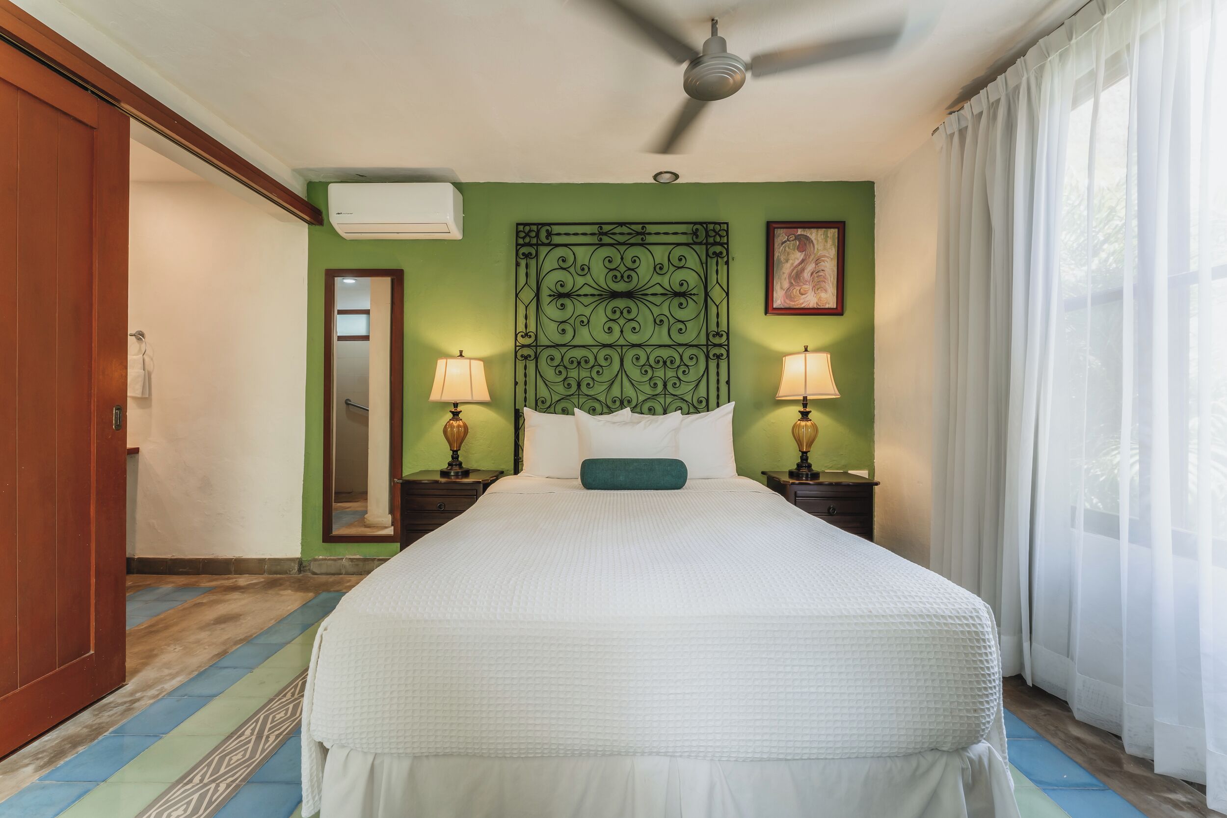 A hotel room with a bed, two bedside lamps, a ceiling fan, a green decorative wall panel, and a window with curtains.