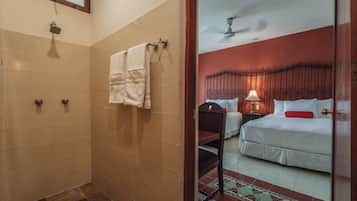 A hotel room with a shower, a bed with white bedding and a red pillow, a nightstand with a lamp, and a patterned rug.