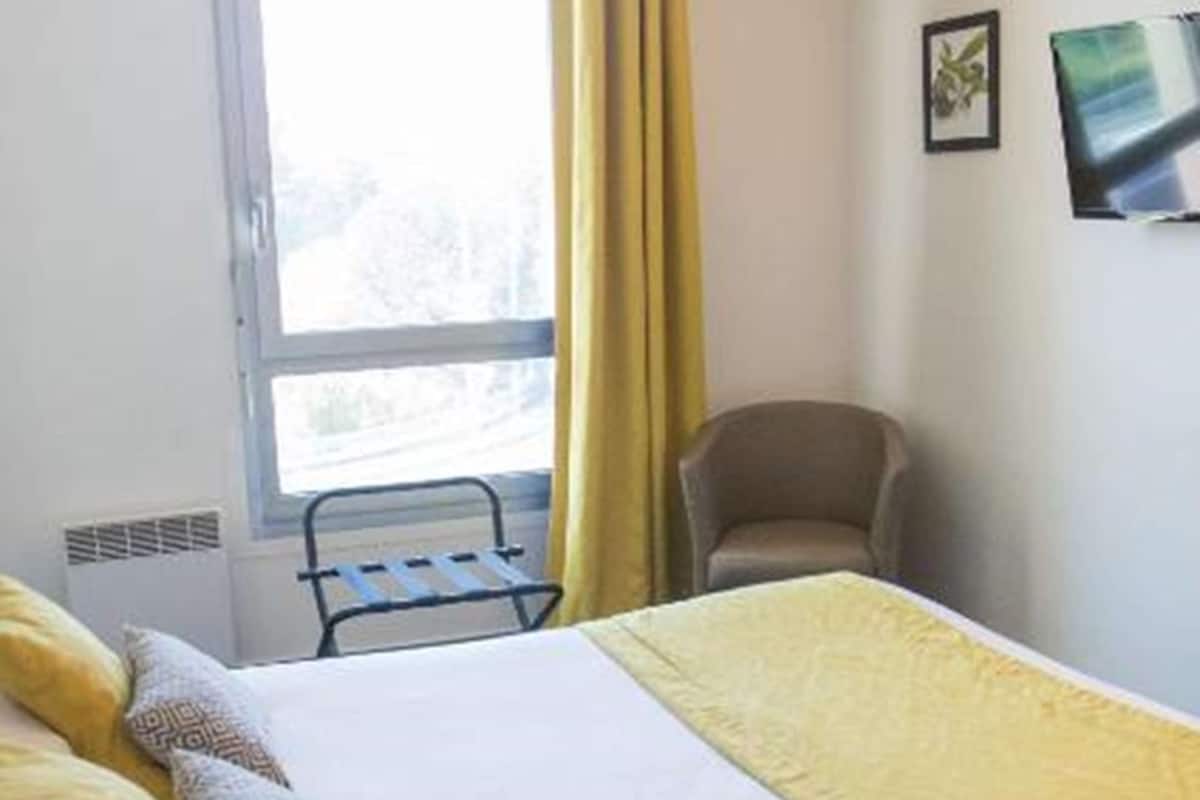 Ein Hotelzimmer mit Bett, Stuhl, Fenster mit gelben Vorhängen und einem an der Wand befestigten Fernseher.