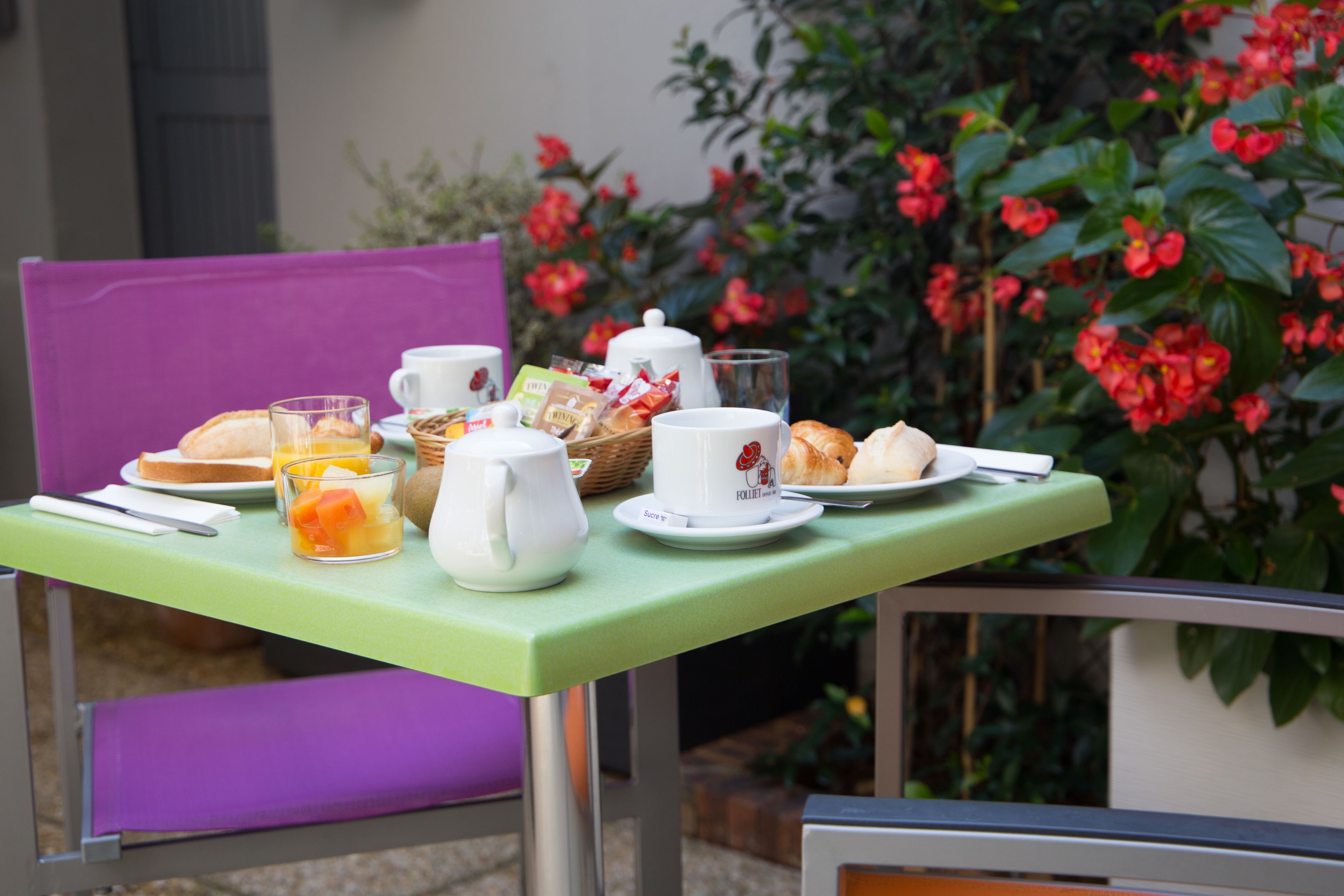 A table set with breakfast items including toast, fruit, and pastries, with a view of red flowers in the background.