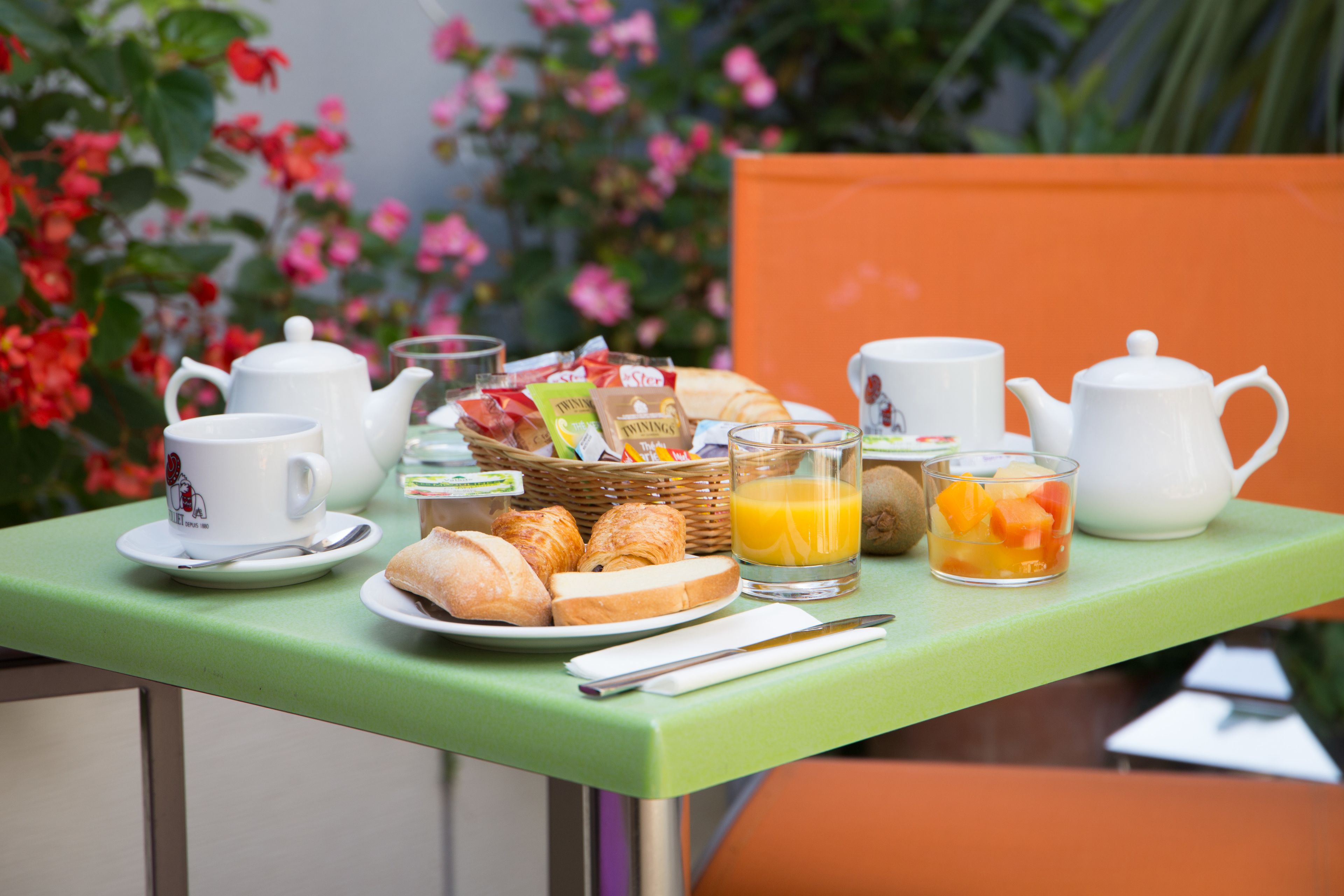 Una mesa de desayuno con mantel verde, tetera blanca, tazas, una cesta de pasteles y un vaso de jugo de naranja.