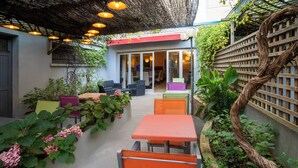 Un espace extérieur aménagé en terrasse, avec des tables et des chaises, un coin salon abrité et une vue sur l’intérieur à travers des portes vitrées.