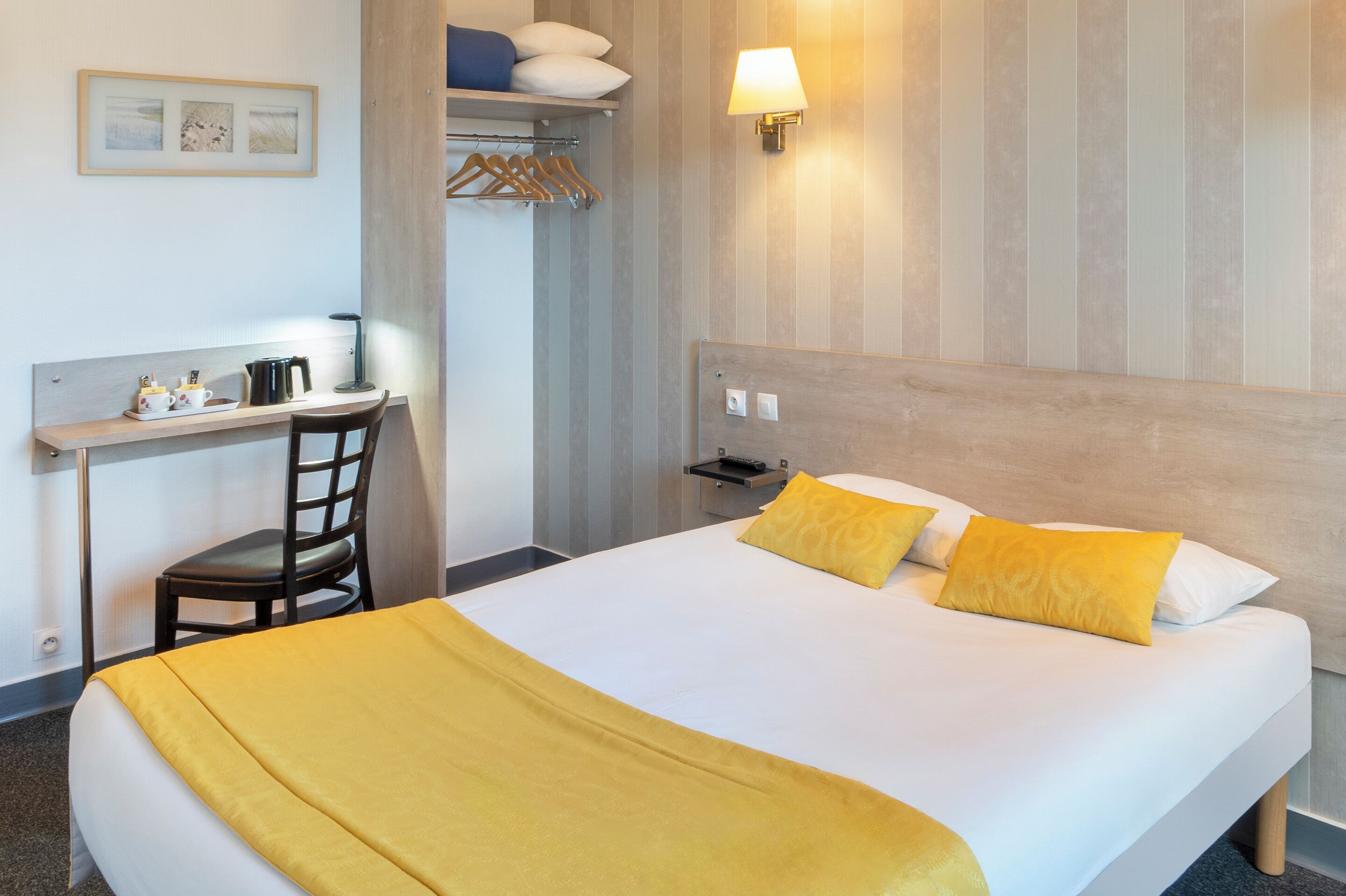 A hotel room with a bed, a desk, a chair, a lamp, and a wall-mounted shelf.