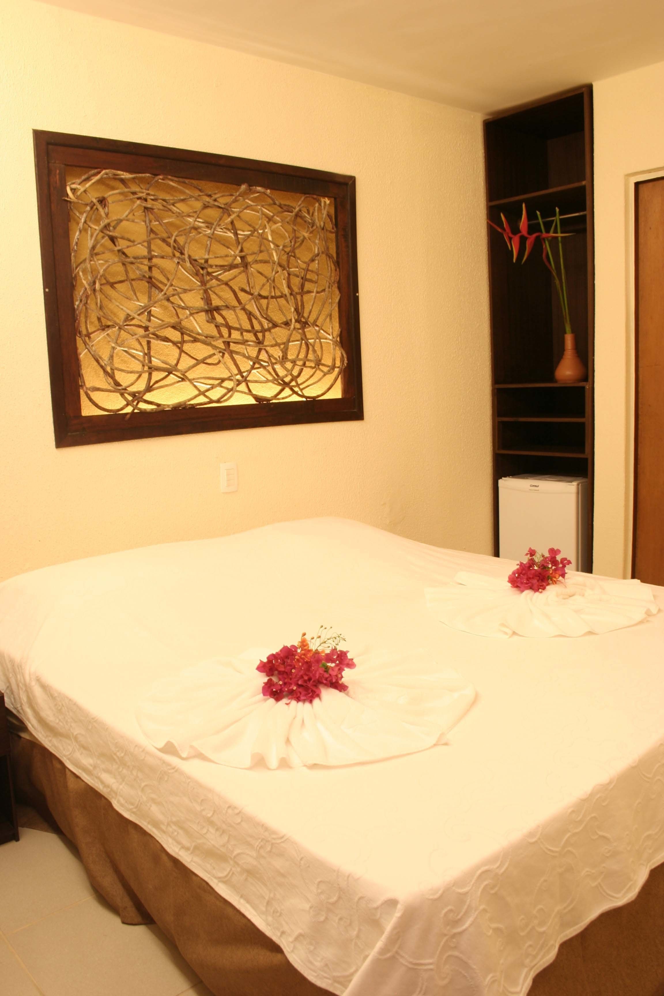 Una cama bien tendida con sábanas blancas y flores decorativas, una obra de arte abstracta colgada en la pared, y un pequeño florero sobre una estantería.
