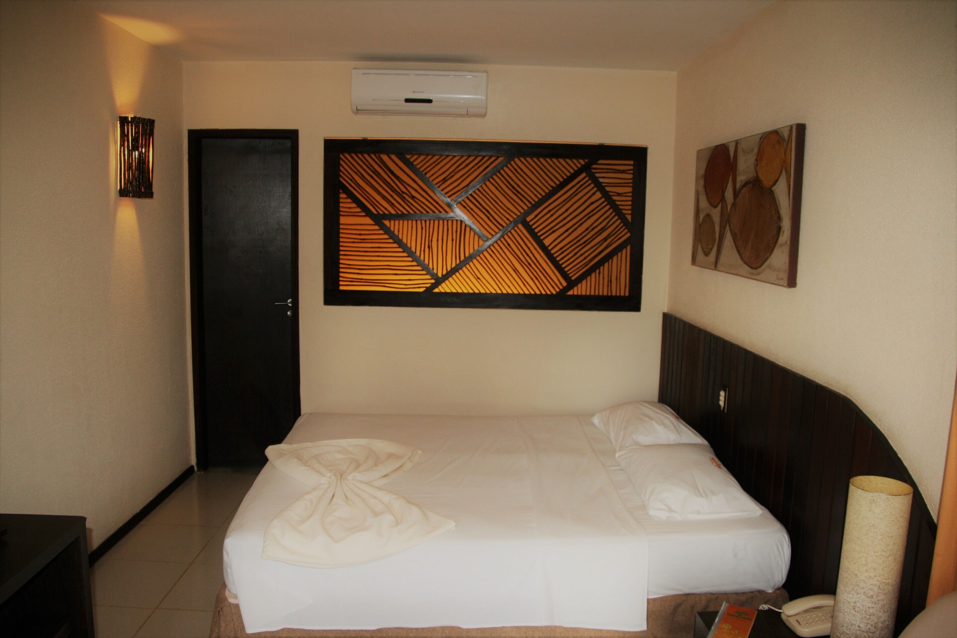 A hotel room with a bed, a wall-mounted air conditioner, a door, and a wall decoration.