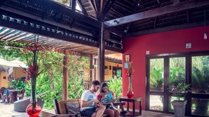 A couple sitting on a bench in a covered outdoor area with a tiled floor, surrounded by potted plants and a red wall. - Hotel Aconchego Porto de Galinhas (Ipojuca)