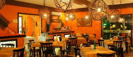 A restaurant interior with orange walls, multiple tables with yellow tablecloths, and hanging spherical light fixtures.
