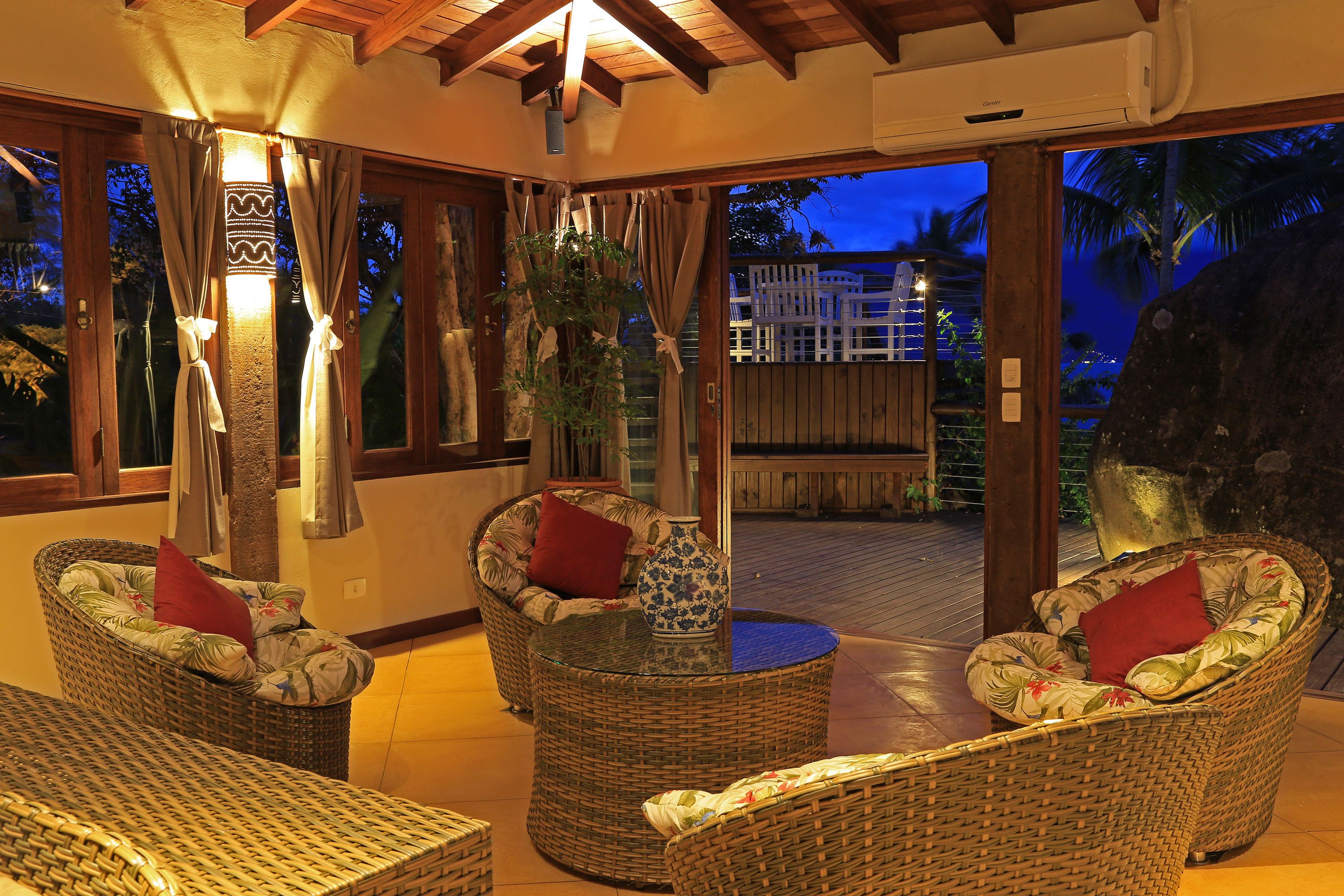 A cozy indoor seating area with wicker furniture and a glass-top table, featuring a decorative vase and a red pillow.