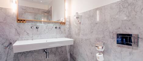 A modern bathroom with a marble wall, a large mirror, a white sink, a towel dispenser, and a showerhead.