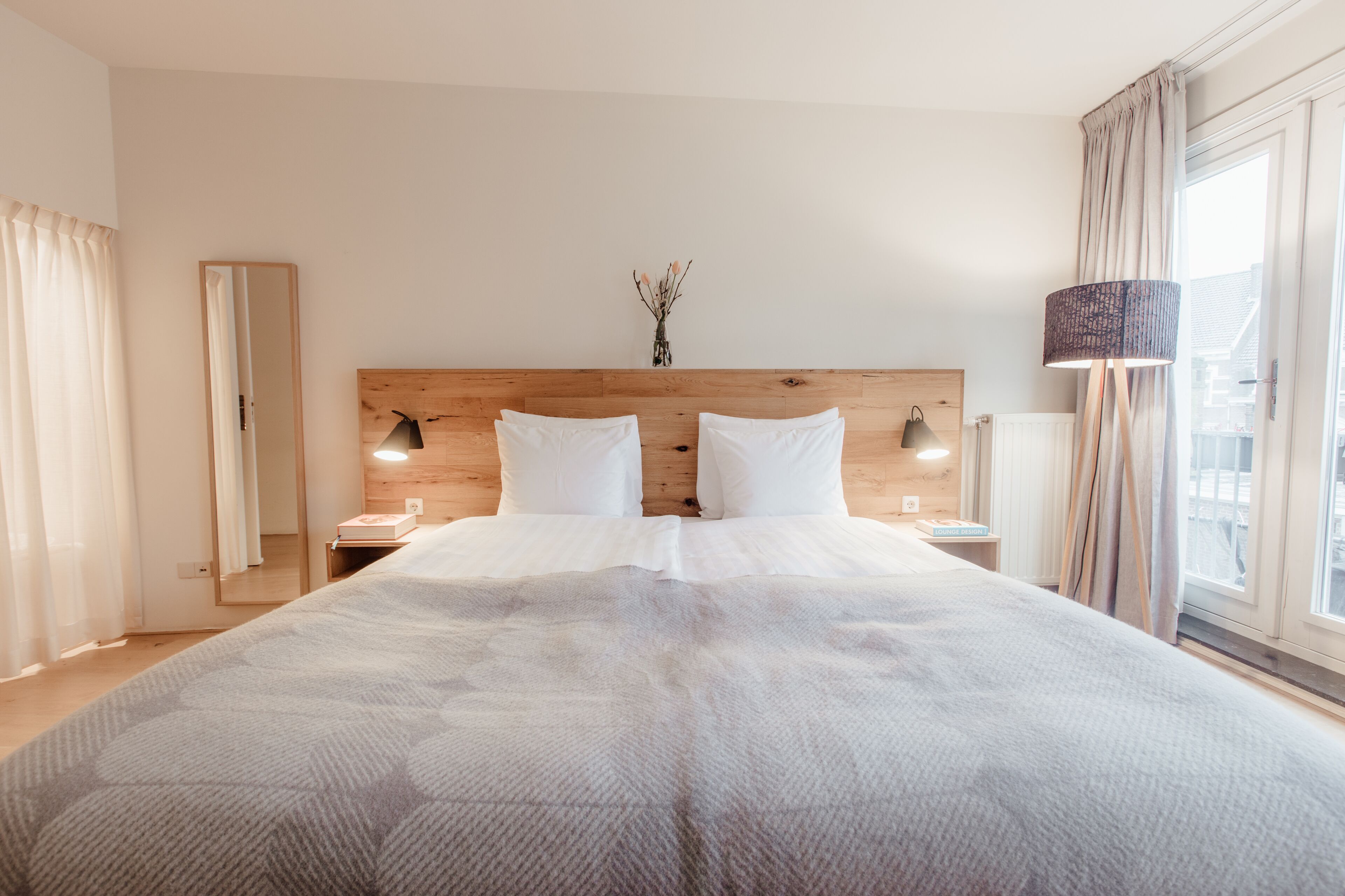 A large bed with a wooden headboard, two bedside tables with lamps, a vase with flowers, and a window with curtains.
