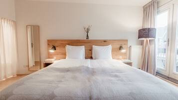 A large bed with a wooden headboard, two bedside tables with lamps, a vase with flowers, and a window with curtains.