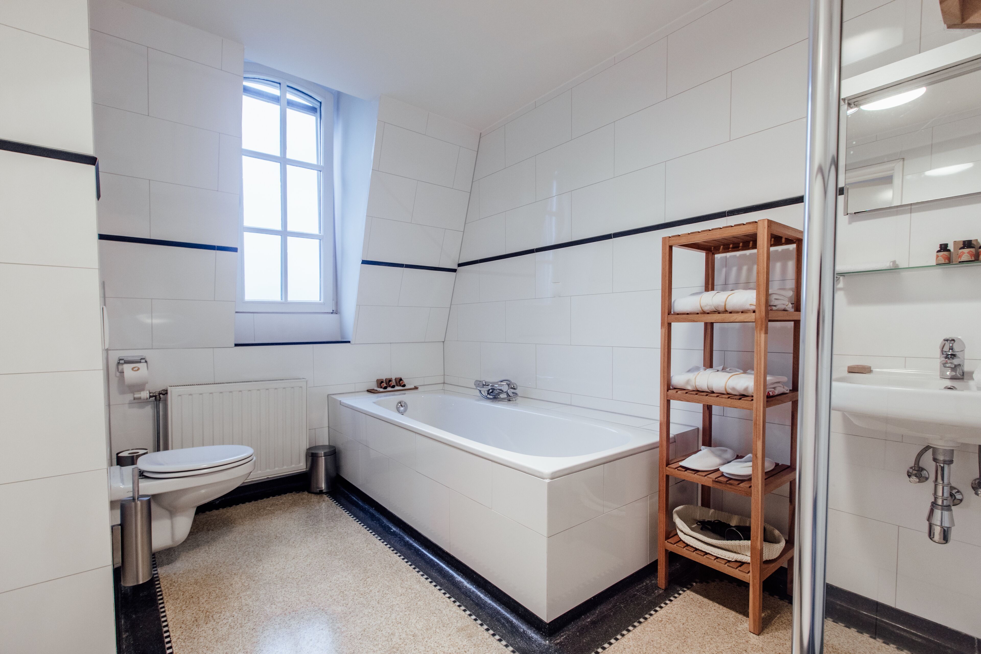 A modern bathroom with a bathtub, a toilet, a window, and a wooden shelf with towels.