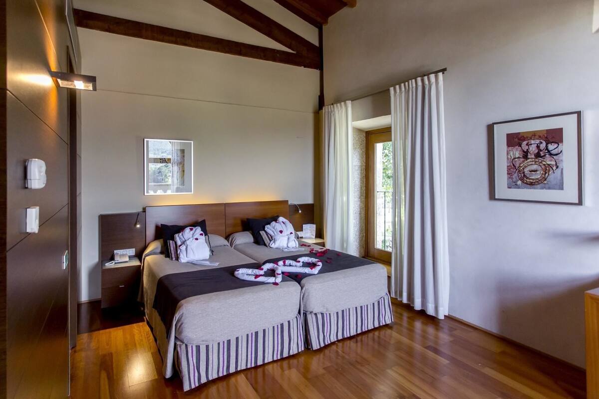 A modern hotel room with a wooden floor, a large bed with a headboard, a bedside table, a nightstand, a framed picture on the wall, and a window with curtains.