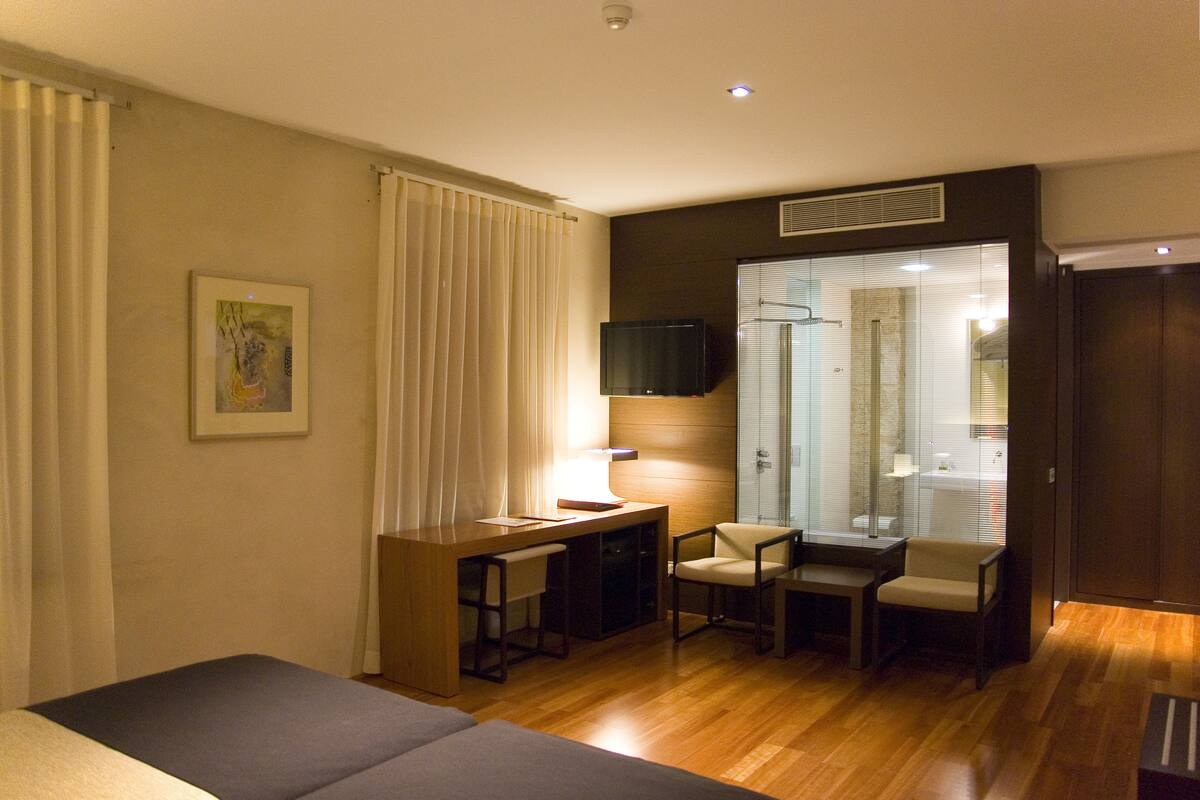 A hotel room with a bed, a desk with a chair, a TV, and a bathroom visible through a glass door.