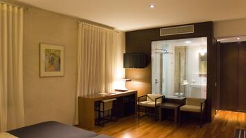 Una habitación de hotel con una cama, un escritorio con silla, un televisor y un baño visible a través de una puerta de cristal.
