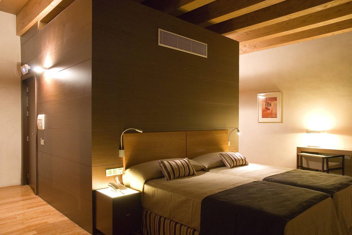 A hotel room with a wooden ceiling, a bed with striped bedding, a nightstand with a lamp, and a small table with a chair.