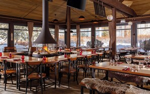 Un restaurante con mesas y sillas de madera, un gran pozo de fuego y amplias ventanas que ofrecen vistas a árboles y nieve.