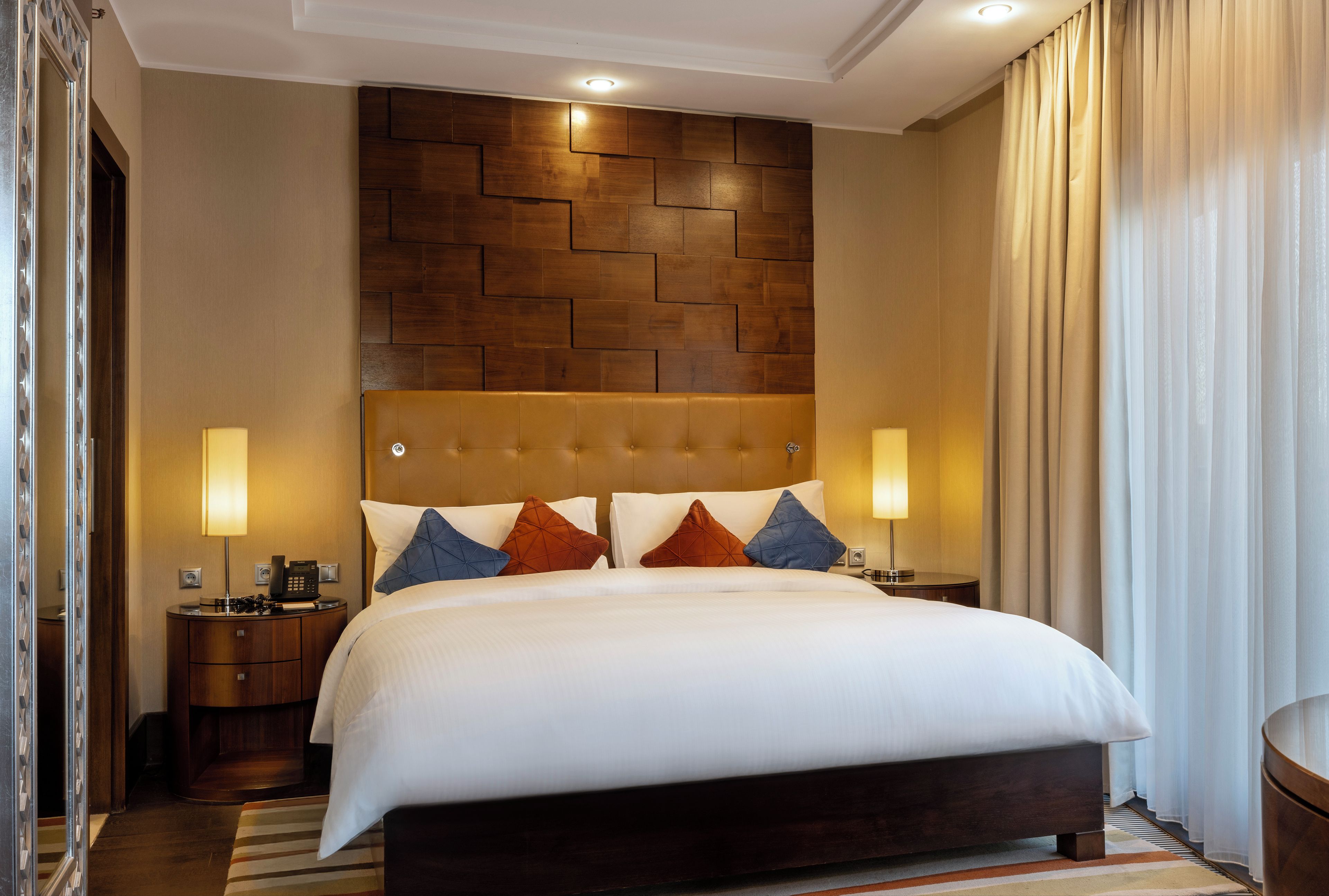 A hotel room with a large bed, a wooden headboard, two bedside lamps, and a view of a curtain.