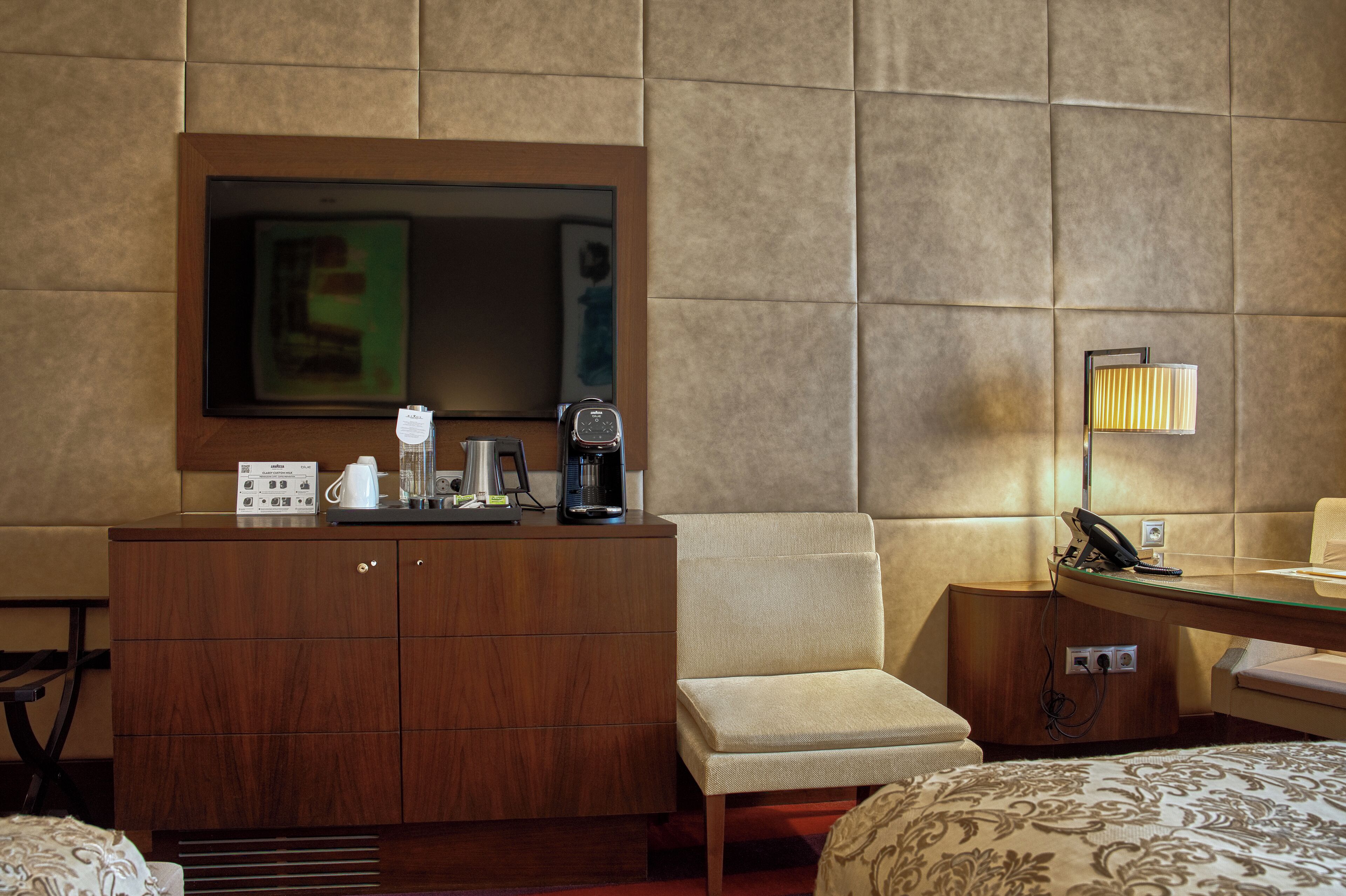 Habitación de hotel con un armario de madera, una pequeña mesa con cafetera, una silla, una cama con ropa de cama estampada y un televisor de pared.