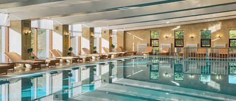 Una amplia piscina interior con varias zonas de descanso y tumbonas, rodeada de grandes ventanales y un muro de azulejos.
