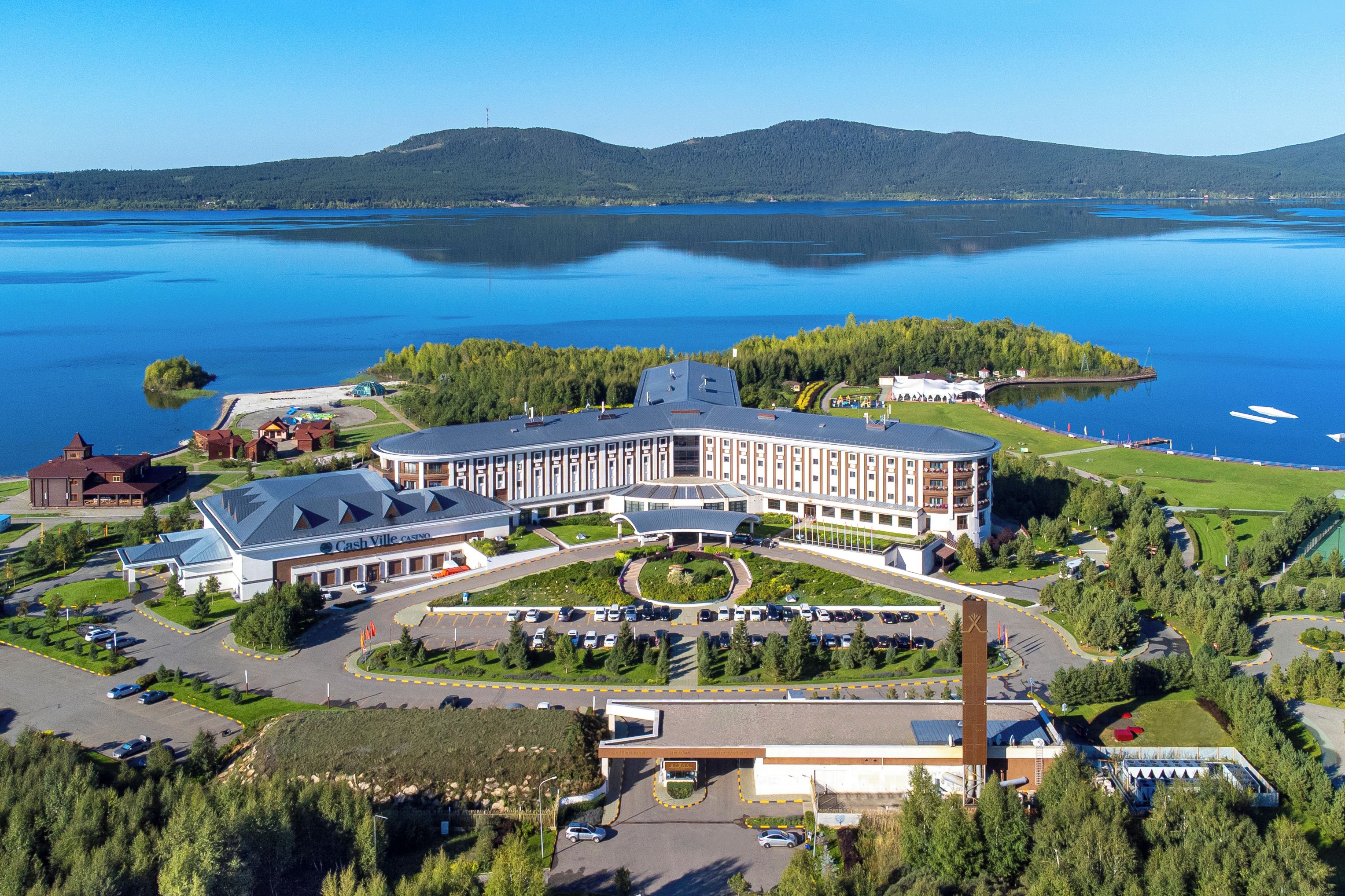 Ein großes Hotel mit einem zentralen Innenhof, umgeben von einem See und bewaldeten Hügeln.