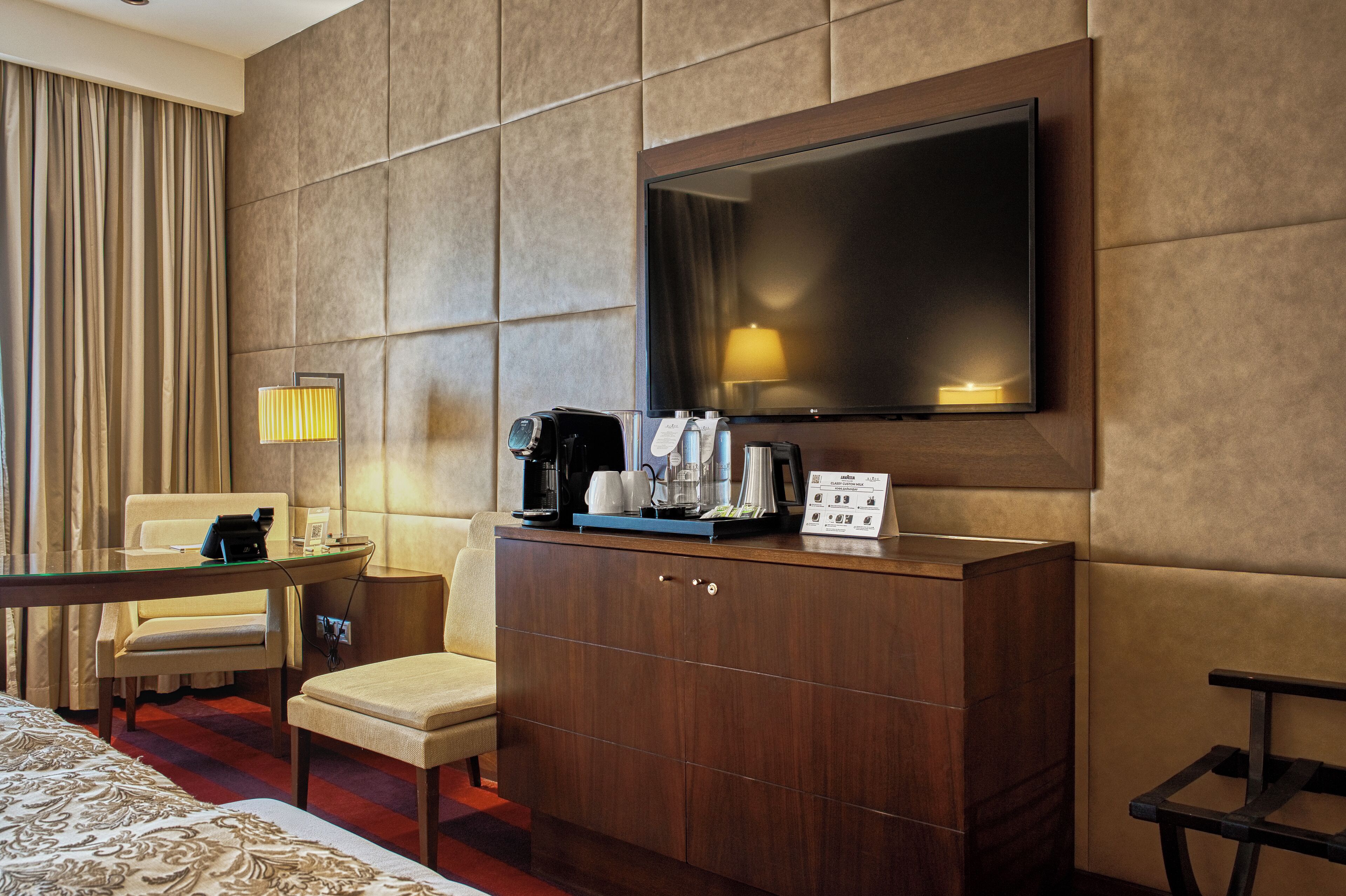 Une chambre d’hôtel dotée d’un grand téléviseur à écran plat fixé au mur, d’un meuble en bois contenant une machine à café, d’une lampe et d’une petite table avec une chaise.